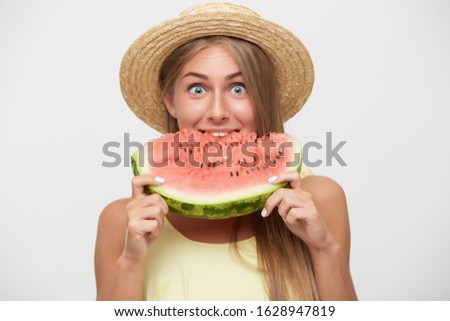 Suprised young pretty cheerful long haired female rounding her blue eyes while looking positively at camera, dressed in boater hat and yellow top while standing over white background