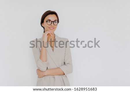 Elegant brunette businesswoman stands thoughtfully indoor, wears formal clothes, big spectacles, keeps hand under chin, stands isolated on white background, copy space for your promotion aside