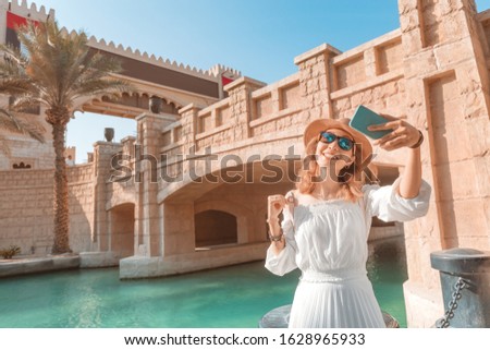 Happy asian girl in white dress taking selfie photo on her smartphone against background of middle eastern old town. Travel and vacation in Dubai and Arab Emirates concept