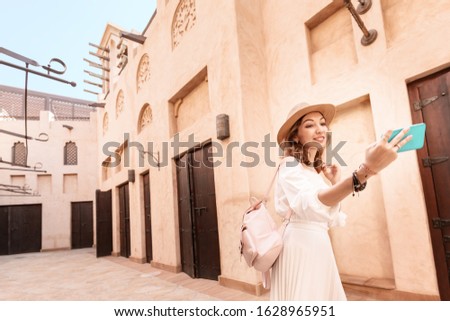 Happy asian girl in white dress taking selfie against narrow streets of old Arabian town in Dubai. Travel destinations and tourism in UAE concept