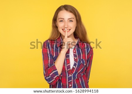 Please, be quiet! Portrait of cheerful ginger girl holding finger on lips making hush silence gesture and smiling, asking to keep secret, don't speak. indoor studio shot isolated on yellow background