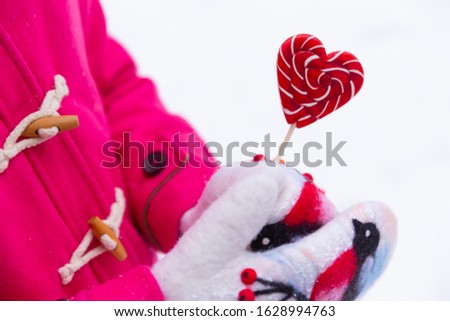 Hands in white felted wool mittens with a bullfinch picture on them hold a heart shaped lollipop. Outdoor Valentine's day concept with copy space.