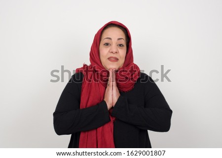 Horizontal portrait of pretty muslim woman keeps palms together, has pleased expression. Glad attractive female makes request, pleads for mercy. Hopeful young adult.