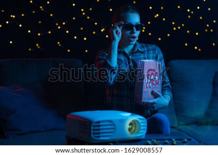 girl in 3D glasses with popcorn on a sofa at home watching movies through a projector