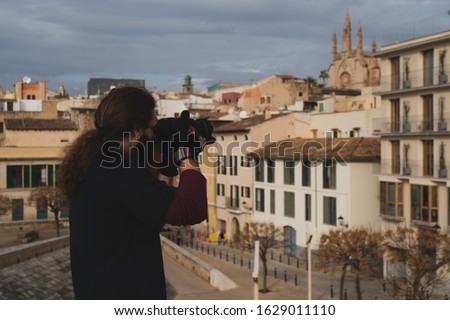 Man with long hair taking a picture with a reflex camera of some buildings and a church from a high terrace