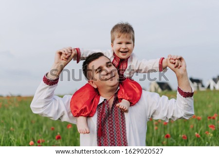 Happiness Smiling Child Son on Father Shoulders. Joyful Funny Characters Wearing Ukrainian National Cossack Garments. Red Flowers Poppy Field, Cloudy Sky and Village on Blurred Background
