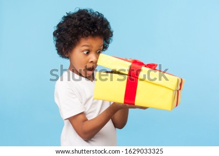 Portrait of amazed curious adorable little boy peeking inside gift box, unpacking present with funny astonished expression, impatient child unboxing birthday surprise. studio shot blue background