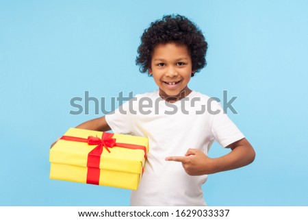 Look at my present! Cheerful amazing cute little boy pointing to gift box and looking at camera with toothy smile, child satisfied with birthday surprise. studio shot isolated on blue background