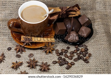 On the table is a cup of coffee with milk and a saucer with chocolates. Next to them are star anise, coffee beans and cinnamon sticks.
