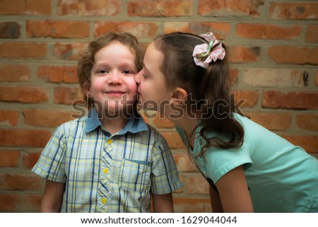 A little girl kisses a boy on the cheek. Brother and sister. Against a brick wall.