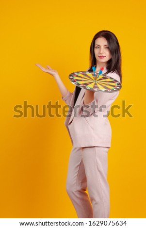 Young brunette girl in a trouser suit with a dartboard in hand on a yellow background