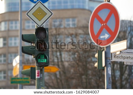 a green traffic light in the city of Berlin with a sticker with the text go vegan, in the background a building and trees 