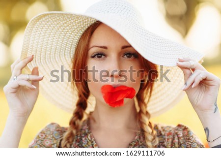 Portrait of beautiful redhead woman in white hat on green field with poppy flower in her mouth in summer sunset