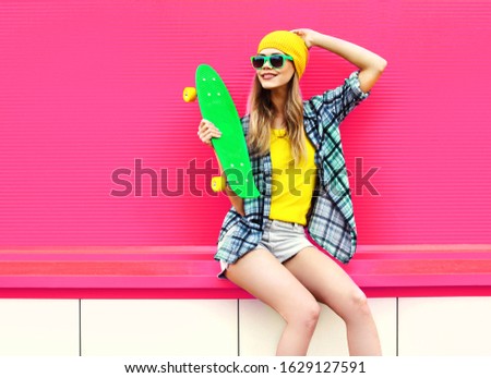 cool smiling woman with skateboard wearing colorful yellow hat on pink background