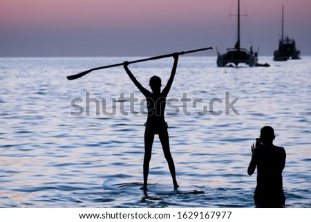 Silhouette of woman stand on surfboard in the sea and lift a paddle in the air and man take a photo for her, Phuket, Thailand