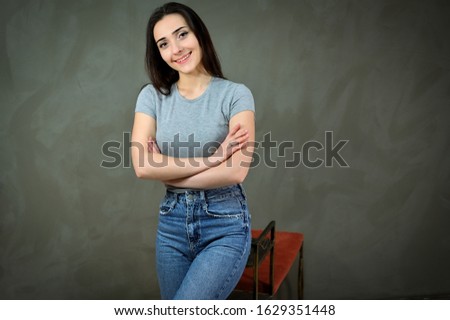 Portrait of a pretty young brunette woman in a gray t-shirt and blue jeans sits on a stand. The concept of a cute girl on a gray background. Smiling, showing hands with emotions.
