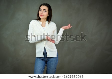 The concept of a cute girl on a gray background. Portrait of a pretty young brunette woman in a white sweater and blue jeans stands near a gray wall. Smiling, showing hands with emotions.