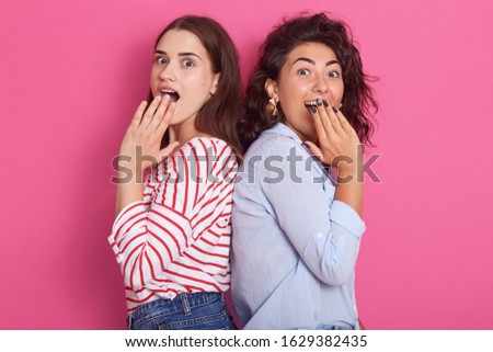 Picture of charismatic curious good looking cute sisters standing back to back, covering their mouths with hands, being impressed by news, having communication time. People and emotions concept.
