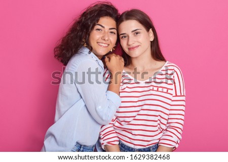 Horizontal picture of two charming sisters standing isolated over pink background in studio, posing close to each other, having pleasant facial expression, enjoying leisure time. Family concept.