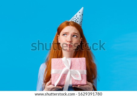 Perplexed and indecisive, redhead girl want open present, thinking with troubled expression concerned, holding pink cute box of gift, wearing birthday cap, look sideways, blue background