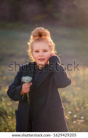 a curly haired blonde girl with a tuft of hair in the background light blows a dandelion in a coat in a meadow in the autumn