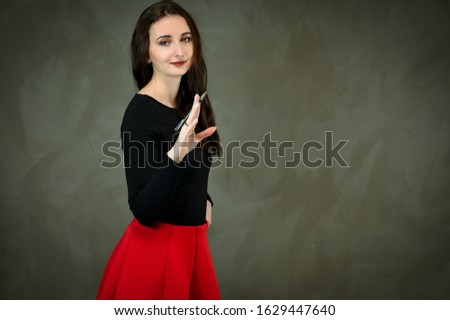 Photo of a pretty slender young brunette woman with good makeup on a gray background. Model in a dark blouse and a red skirt. Talking and showing hands with a smile