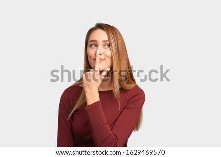 Funny girl holding index finger on nose, smiling sweetly. Beautiful young woman with long hair, clean smooth skin isolated on white background in Studio with an empty space for text and advertising. Royalty-Free Stock Photo #1629469570