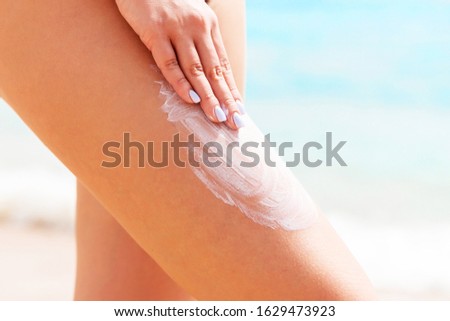 Woman in a black swimsuit is applying sun protection with her fingers on her leg at the beach.