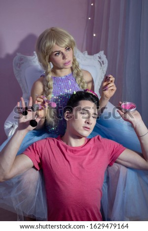 A young beautiful woman in the image of a doll in a lilac dress with a blue skirt holds makeup brushes in her hand in front of her is a young man with makeup in a bright pink t-shirt and holds makeup 