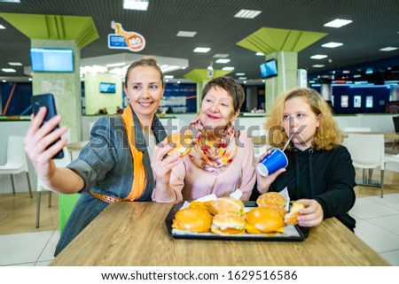 women of different ages are sitting in a fast food restaurant, ordered hamburgers and take selfies. Granny, a woman and a child take pictures. people have fun