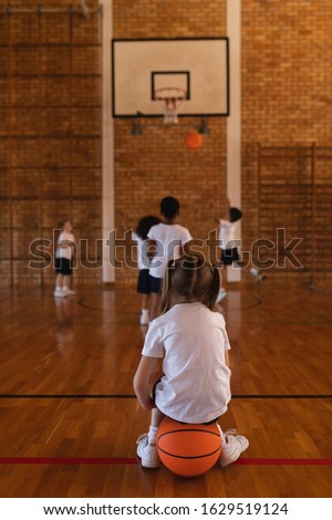 Rear view of schoolgirl sitting on basketball at basketball court in school