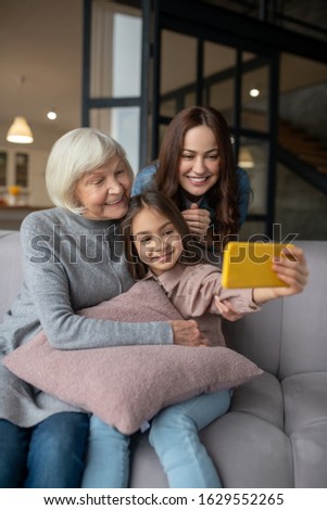 Family photo. Cheerful little girl taking a selfie with her grandmother hugging her on the couch and mom standing behind the couch and leaning forward, all beautiful and happy.