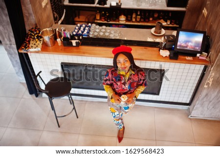 Enthusiastic african american woman in trendy coloured outfit with red beret chilling in cozy cafe, standing near bar counter.