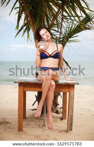 Caucasian woman in bikini siting on wooden table near palm tree on beach and drink Pina Colada 