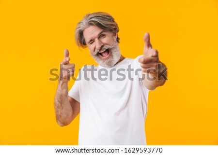 Image of emotional cheerful senior grey-haired bearded man posing isolated over yellow wall background pointing to you.