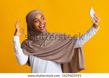 Cheerful black muslim girl in hijab taking selfie on smartphone and showing ok gesture over yellow background with free space
