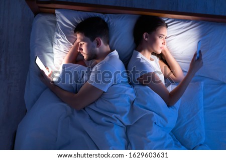 Infidelity. Cheating Spouses Using Smartphones Texting With Lovers Lying Back-To-Back In Bed At Home At Night. Top View, Low-Light Royalty-Free Stock Photo #1629603631