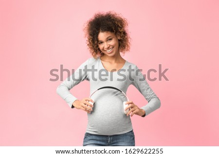 Pregnancy therapy. Afro woman holding headphones on her belly, pink background