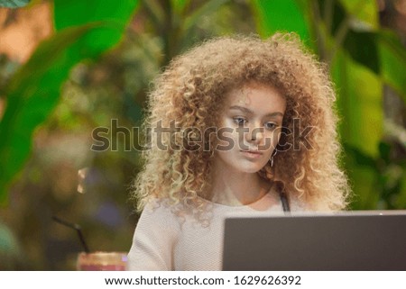 Young businesswoman with curly hair working on laptop computer at the table with green plants in the background