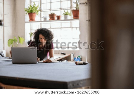 Young woman writing notes and working on a laptop while sitting at a workbench in her picture framing studio