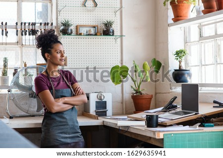 Confident young woman looking out of a window while leaning with her arms crossed against a workbench in her picture framing studio