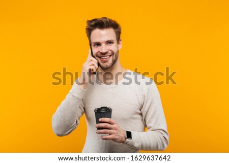 Attractive happy young man wearing pullower standing isolated over yellow background, talking on mobile phone while drinking takeaway coffee cup