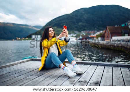 The girl tourist takes a photo on the phone by the lake in Norway. Young woman takes selfie against the backdrop of the mountains in the Norway. Travelling, lifestyle, adventure, concept.