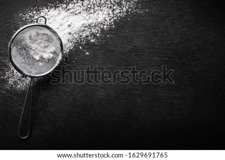 Powdered sugar scattered on a black stone countertop with a sifter for sifting , top view.