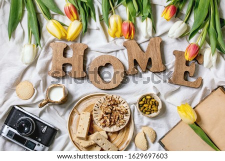 Wooden decorative letters word HOME with colorful tulips, different snacks, milk jug, cookies, vintage book and camera over folded cotton cloth. Flat lay, space. Spring home decoration.