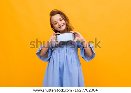 cute smiling teen in a blue dress shows the phone in horizontal position with a mockup on a yellow background with copy space