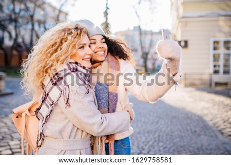 Always online concept.Emotional portrait of excited african female hugging, talking selfie portrait with girl friend outdoors in city street.Curly-hair biracial friends in scarfes in sunny winter day