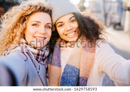 in the city, two female multiethnic curly friends traveling together, she takes the opportunity to make a selfie portrait with a smartphone,old street city on background. Sun glare effect.