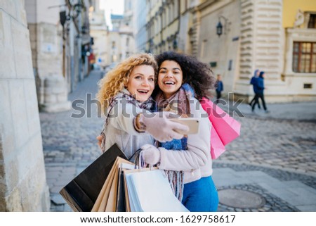 Two beautiful smiling multiethnic sisters, many colorful bags are taking selfie in the shopping time at Christmas holiday in the city center.
