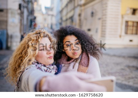 Outdoor close up portrait of two curly blond and brunette multiethnic happy smiling girls making selfie on street.City lifestyle. Ladies wearing stylish winter coats, jacket. City street on background